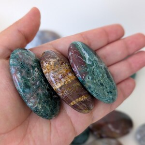 K&ouml;nnte beinhalten: Drei polierte Edelsteine in Gr&uuml;n-, Braun- und Burgundert&ouml;nen werden in einer Hand gehalten. Die Steine haben eine ovale Form und eine gl&auml;nzende Oberfl&auml;che. Die Edelsteine sind glatt und scheinen Natursteine zu sein.