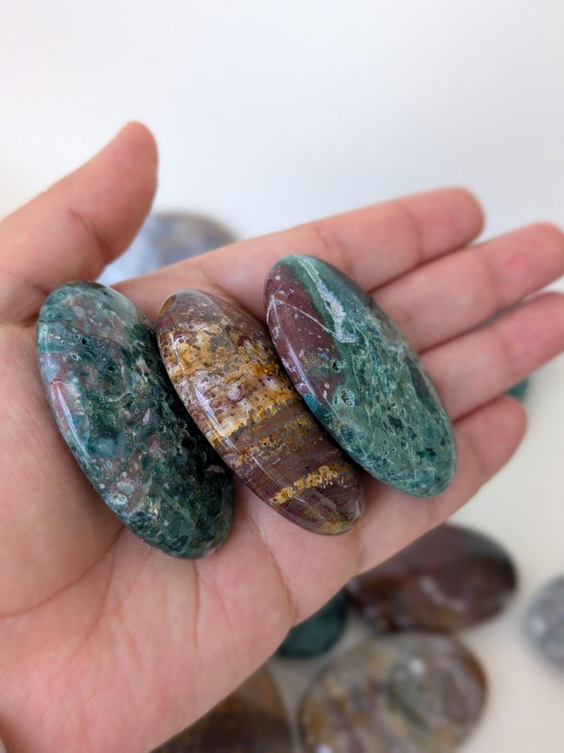 K&ouml;nnte beinhalten: Drei polierte Edelsteine in Gr&uuml;n-, Braun- und Burgundert&ouml;nen werden in einer Hand gehalten. Die Steine haben eine ovale Form und eine gl&auml;nzende Oberfl&auml;che. Die Edelsteine sind glatt und scheinen Natursteine zu sein.