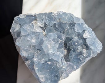 May include: A cluster of pale blue celestite crystals. The crystals have a rough, textured surface and are arranged in a geode-like formation. The stone is resting on a white and grey marble surface.