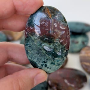 K&ouml;nnte beinhalten: Ein oval geschliffener Edelstein mit einer Mischung aus tiefroten, gr&uuml;nen und braunen Farben. Der Stein wird in einer Hand gehalten, im Hintergrund sind weitere &auml;hnliche Steine zu sehen. Der Edelstein hat eine glatte, reflektierende Oberfl&auml;che.