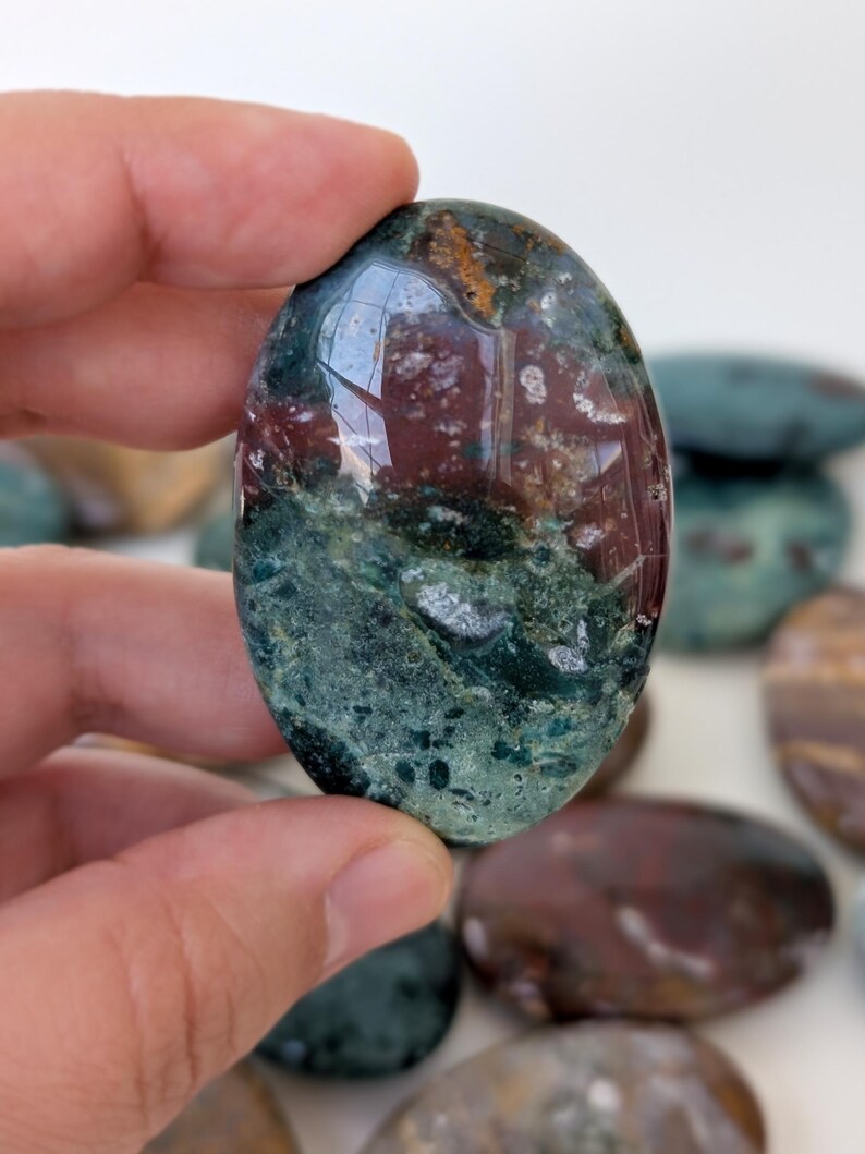 K&ouml;nnte beinhalten: Ein oval geschliffener Edelstein mit einer Mischung aus tiefroten, gr&uuml;nen und braunen Farben. Der Stein wird in einer Hand gehalten, im Hintergrund sind weitere &auml;hnliche Steine zu sehen. Der Edelstein hat eine glatte, reflektierende Oberfl&auml;che.