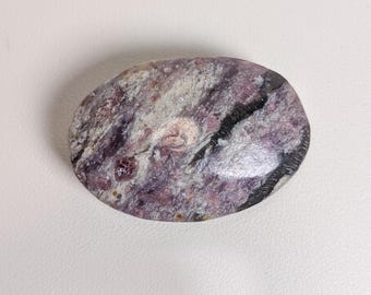 May include: An oval-shaped polished gemstone featuring a mottled pattern of deep purple, lavender, and white mineral inclusions with a dark black streak running across the right side of the smooth, reflective surface of the stone.