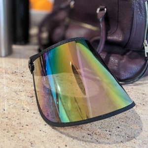 May include: A visor with a black frame and an iridescent, reflective shield. The shield displays a range of colours, including green, yellow, and blue. The visor is on a speckled surface, with a purple handbag in the background.