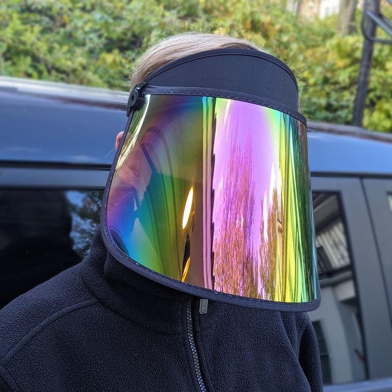 May include: A black visor featuring a large, iridescent shield. The shield reflects a spectrum of colours, including green, blue, and purple. The visor is worn by a person wearing a black fleece jacket.