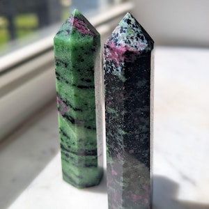 May include: Two polished hexagonal crystal towers standing upright. The left tower is primarily bright green with black streaks and magenta inclusions. The right tower is darker, featuring a mix of deep green, black, and magenta mineral deposits.