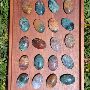 K&ouml;nnte beinhalten: Ein Holztablett mit neunzehn polierten, ovalen Edelsteinen. Die Steine zeigen eine Vielzahl von Farben, darunter Gr&uuml;n, Braun, Rot und Beige. Die Edelsteine sind in einem Raster angeordnet.