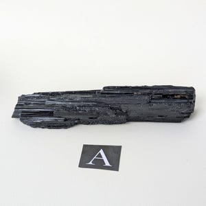 May include: A black tourmaline crystal with a rough, textured surface. The crystal is long and rectangular, with visible striations. A small square card with the letter "A" is next to the crystal.