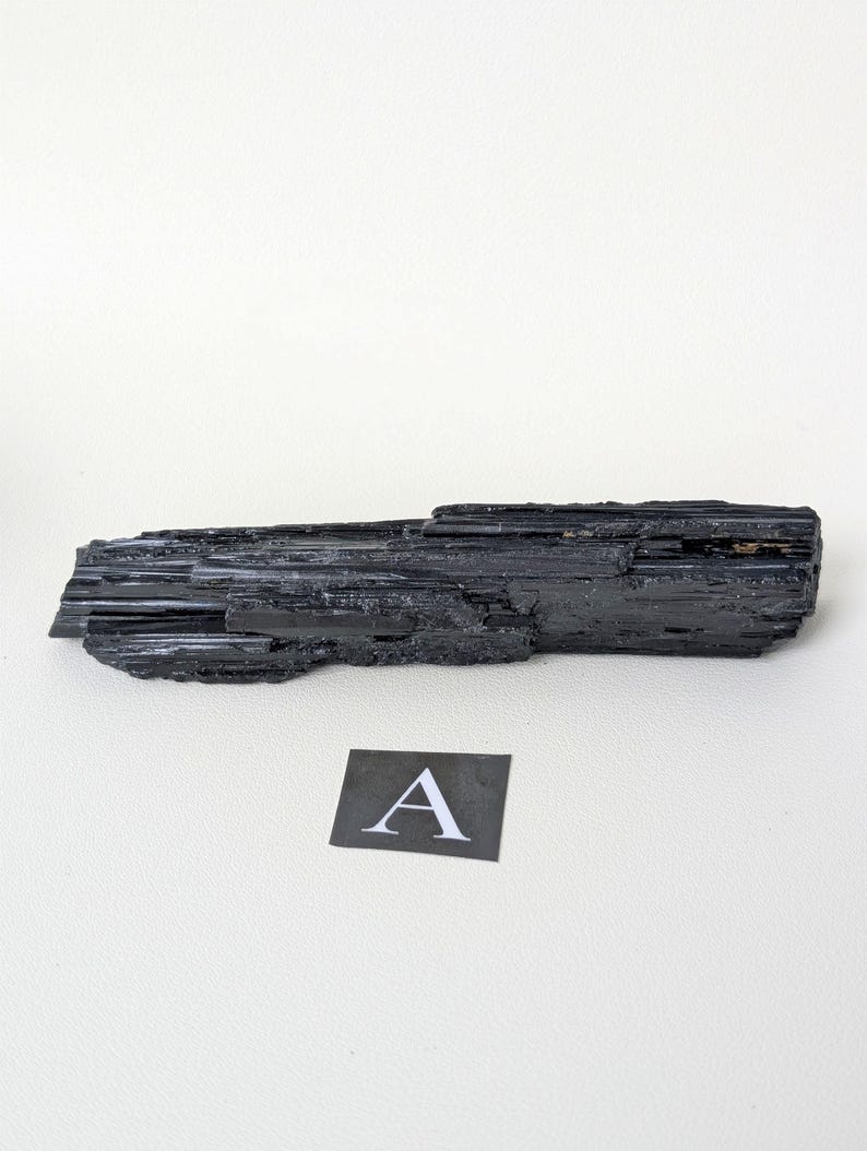 May include: A black tourmaline crystal with a rough, textured surface. The crystal is long and rectangular, with visible striations. A small square card with the letter "A" is next to the crystal.