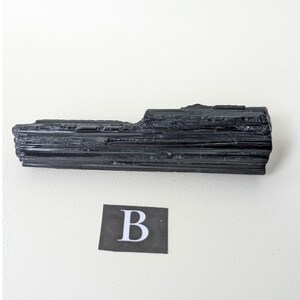 May include: A black tourmaline crystal specimen with a rough, textured surface. The crystal is a dark, solid black color and has a rectangular shape. A small black square with the letter "B" is in the lower portion of the image.
