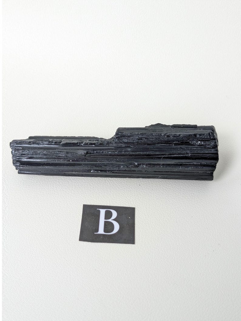 May include: A black tourmaline crystal specimen with a rough, textured surface. The crystal is a dark, solid black color and has a rectangular shape. A small black square with the letter "B" is in the lower portion of the image.