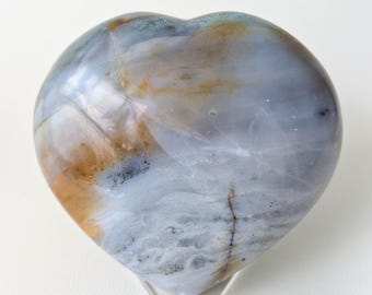 May include: A heart-shaped stone sculpture with a polished surface. The stone features a blend of blue, white, and brown hues, with a translucent quality. The sculpture is displayed on a clear stand, showcasing its natural beauty.