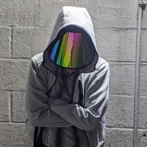 May include: A person in a grey hooded jacket with a rainbow-coloured reflective visor. The jacket has a zip and a black and white patterned shirt. The person's arms are crossed. The background is a grey brick wall.