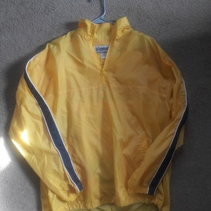 Puede incluir: Chaqueta cortavientos amarilla con media cremallera y dobladillo con cordón. Las mangas tienen rayas negras y blancas. La etiqueta dice "AEROPOSTALE". La chaqueta está colgada de una percha plateada.