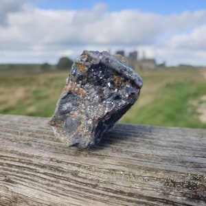 May include: A close-up of a dark grey rock with black crystals embedded in it. The rock is sitting on a weathered wooden surface.