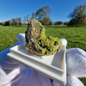 Puede incluir: Primer plano de un espécimen mineral con una roca rugosa de color gris oscuro, cubierta de formaciones cristalinas de color verde brillante y verde amarillento. La muestra se sostiene en una bandeja blanca sobre un fondo de hierba verde y cielo azul.