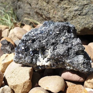 May include: A large, dark gray, metallic rock with a rough, textured surface. The rock is surrounded by smaller, smooth, gray and brown rocks.