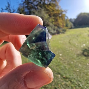May include: A translucent fluorite crystal featuring a geometric cubic structure with distinct colour zoning that transitions from deep violet and indigo at the top to a vibrant emerald green at the base, showing natural internal inclusions.