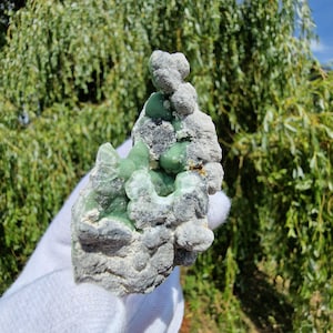 May include: A raw mineral specimen featuring clusters of smooth, pale green botryoidal formations embedded within a rough, textured grey and white matrix. The mineral displays a translucent quality in the green sections, contrasting with the opaque, rocky exterior of the surrounding stone structure.
