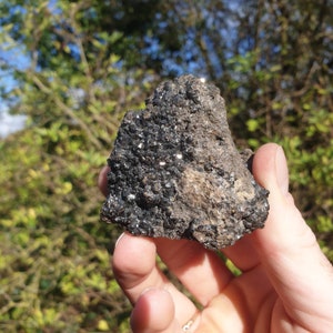 May include: A close-up of a dark gray rock with a rough texture and small, shiny black crystals embedded in it.