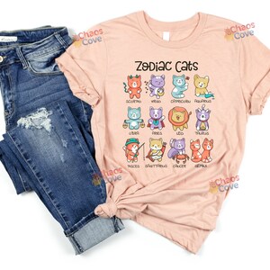May include: A pink t-shirt with the text "Zodiac Cats" and illustrations of cats representing each astrological sign. The cats are in various colors and poses, with each sign's name written below.