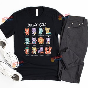 May include: Black t-shirt with a white graphic of zodiac cats. The graphic shows a cat for each zodiac sign with the sign's name underneath. The shirt is laid flat on a white surface with a pair of white sneakers and a pair of dark gray jeans.