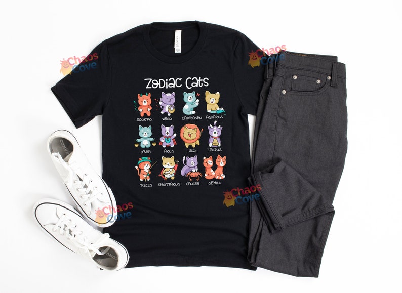 May include: Black t-shirt with a white graphic of zodiac cats. The graphic shows a cat for each zodiac sign with the sign's name underneath. The shirt is laid flat on a white surface with a pair of white sneakers and a pair of dark gray jeans.