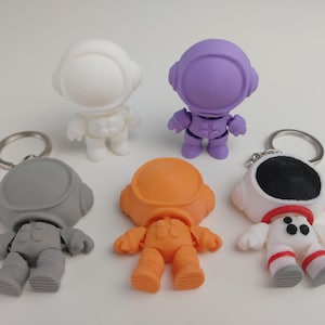 May include: Five astronaut keyrings in different colours: white, purple, grey, orange, and white with black accents. Each keyring features a small astronaut figure with a round helmet.