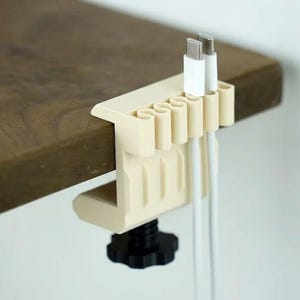 May include: A cream-coloured desk cable organiser with a clamp and a black knob, holding two white USB-C cables. The organiser is attached to a wooden desk. The design features multiple slots for cable management.