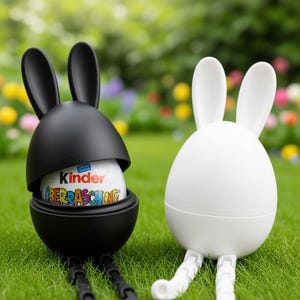 Puede incluir: Dos figuras con forma de conejito, una negra y abierta que contiene un huevo Kinder Sorpresa, la otra blanca y cerrada. Ambas tienen orejas largas y están sobre hierba verde.