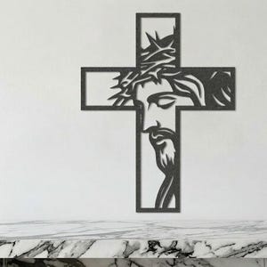 May include: A dark metal cross wall art featuring a detailed depiction of Jesus Christ's face, including a crown of thorns. The cross is mounted on a white wall above a marble countertop. The artwork is a religious home decor item.