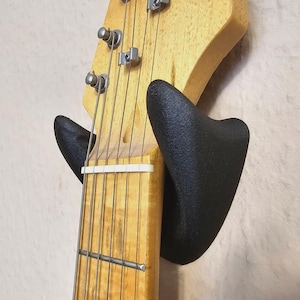 Puede incluir: Un soporte de pared negro para guitarra con una guitarra colgada. La guitarra tiene un acabado de madera natural y una golpeador blanco.