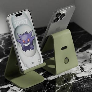 May include: Two smartphones rest on olive green phone stands. One phone displays a purple cartoon character on its screen. The other phone is silver. The stands are on a black and white marble surface.