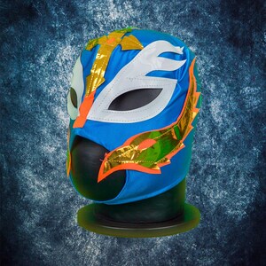 Op de afbeelding: Een levendig blauw worstelmasker met witte, oranje en gouden accenten. Het masker heeft een gestileerd oogontwerp en decoratieve gouden folie-elementen. Het masker wordt gepresenteerd op een zwarte standaard tegen een getextureerde blauwe achtergrond.