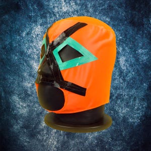 Op de afbeelding: Oranje worstelmasker met zwarte en turquoise accenten. Het masker heeft zwarte oog- en neusopeningen, turquoise driehoeken en zwarte lijnen. Het wordt getoond op een zwarte standaard tegen een blauwe achtergrond.