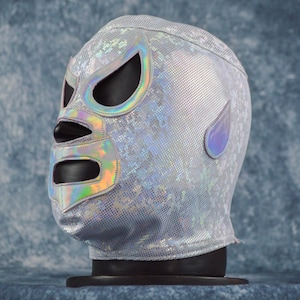 May include: Silver lucha libre mask with iridescent accents around the eye and mouth openings. The mask has a textured, shimmering surface and a teardrop-shaped detail on the side. The mask is displayed on a black stand against a blue background.