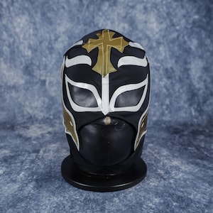 May include: A black wrestling mask featuring white trim around the eye openings and gold accents on the sides. A prominent gold cross is centered on the forehead area, extending toward the top of the head. The mask is displayed on a black stand.