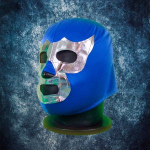 Op de afbeelding: Een blauw lucha libre worstelmasker met een zilverkleurig metallic gezichtsontwerp. Het masker heeft oog- en mondopeningen en wordt gepresenteerd op een groene standaard. Een klassiek worstelaccessoire.