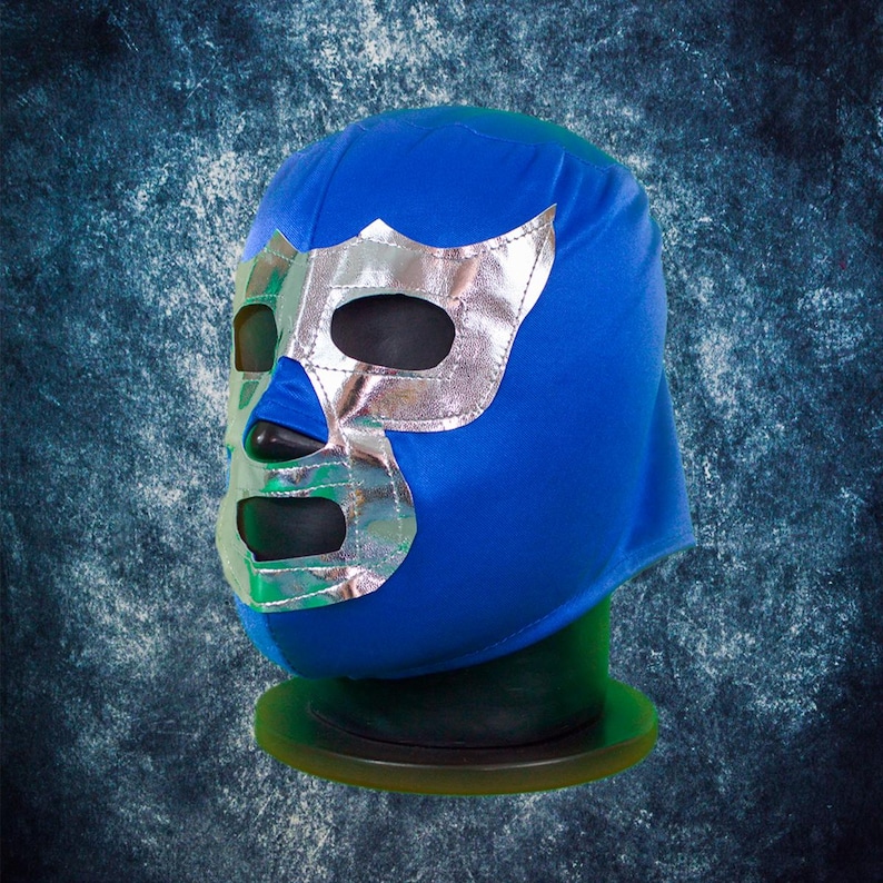 Op de afbeelding: Een blauw lucha libre worstelmasker met een zilverkleurig metallic gezichtsontwerp. Het masker heeft oog- en mondopeningen en wordt gepresenteerd op een groene standaard. Een klassiek worstelaccessoire.