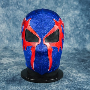 May include: A blue wrestling mask with red accents and black eye coverings. The mask is displayed on a black stand against a textured blue background. The red design features sharp, angular shapes.
