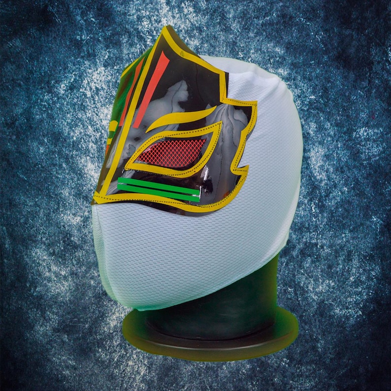 Op de afbeelding: Een wit lucha libre masker met een zwart, geel, rood en groen ontwerp. Het masker heeft een gedetailleerde afbeelding op de voorkant en wordt gepresenteerd op een groene standaard. Het masker is gemaakt van een ademende stof.