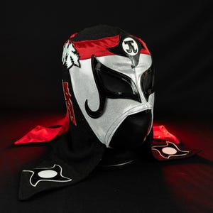 Può includere: Una maschera da wrestling Lucha Libre nera, argentata e rossa. La maschera presenta un volto argentato con dettagli neri per occhi e bocca. Accenti rossi sulla parte superiore e sui lati, con un cappuccio nero ed elementi decorativi.