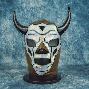 May include: A professional wrestling mask with a bull-like design. The mask features a brown base with silver and black accents, including horns and a stylized face. The mask is displayed on a black stand against a blue background.