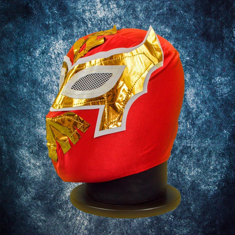 Op de afbeelding: Een rode lucha libre worstelmasker met gouden en witte accenten. Het masker heeft een gouden rand rond de oog- en mondopeningen, met een witte mesh oogbedekking. Het masker wordt getoond op een zwarte standaard tegen een blauwe achtergrond.