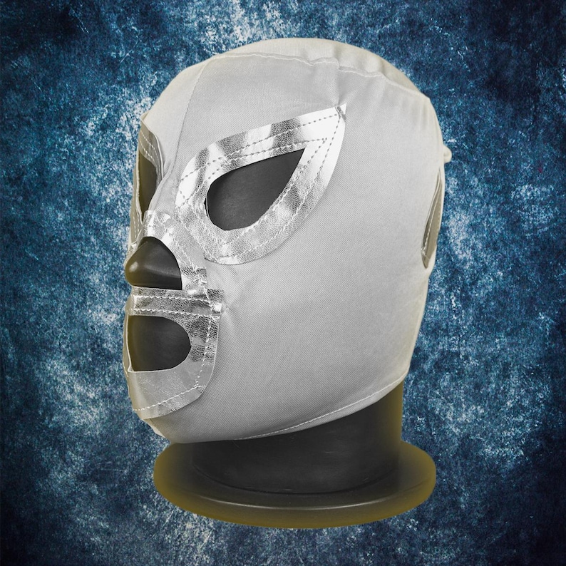 Op de afbeelding: Een zilveren lucha libre worstelmasker met metallic zilveren randen rond de oog- en mondopeningen. Het masker wordt getoond op een zwarte mannequinhoofd tegen een blauwe achtergrond.