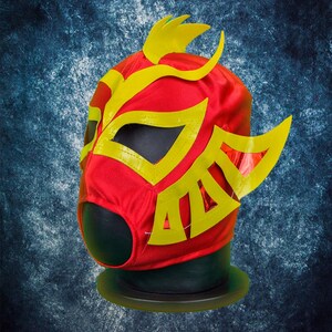 Op de afbeelding: Een felrood worstelmasker met gele accenten. Het masker heeft een gestileerd ontwerp met gele randen rond de oogopeningen en decoratieve vormen. Het wordt getoond op een zwarte mannequinhoofd tegen een getextureerde blauwe achtergrond.