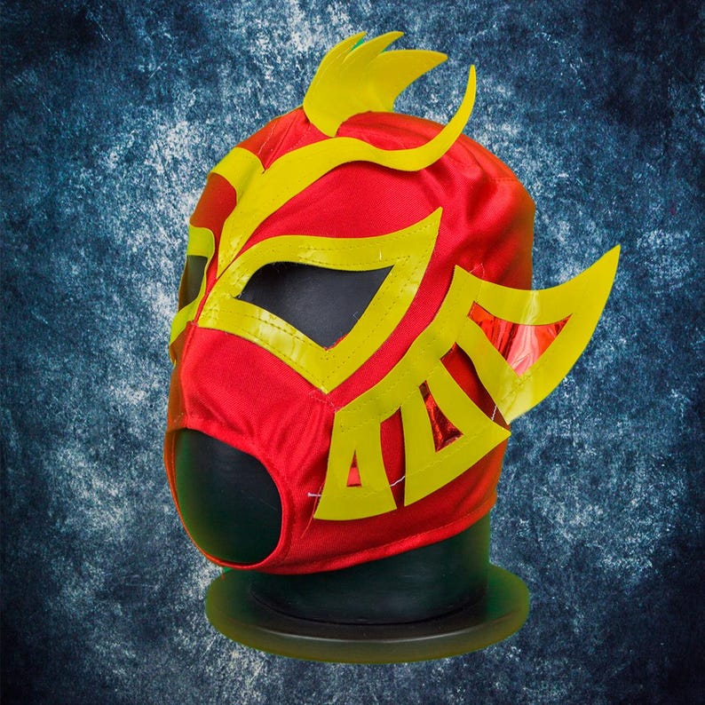 Op de afbeelding: Een felrood worstelmasker met gele accenten. Het masker heeft een gestileerd ontwerp met gele randen rond de oogopeningen en decoratieve vormen. Het wordt getoond op een zwarte mannequinhoofd tegen een getextureerde blauwe achtergrond.