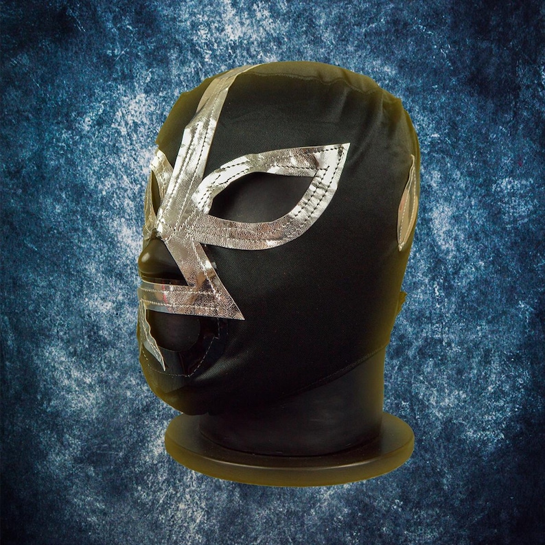 Op de afbeelding: Zwart worstelmasker met zilveren details. Het masker heeft een zilveren ontwerp rond de ogen en over het voorhoofd. Het wordt getoond op een zwarte standaard tegen een blauwe achtergrond.