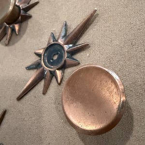 May include: Close-up of several copper-coloured starburst-shaped decorative elements. Each starburst has pointed rays and a central circular depression. A few round, flat copper discs are also visible. The items are arranged on a neutral-toned surface.