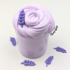 May include: Purple slime in a clear jar with a lavender flower on top. There are several lavender flowers scattered around the jar.