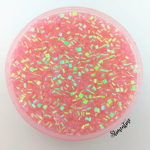 May include: A close-up of a clear container filled with pink iridescent glitter. The glitter is in the shape of small squares and is very sparkly.
